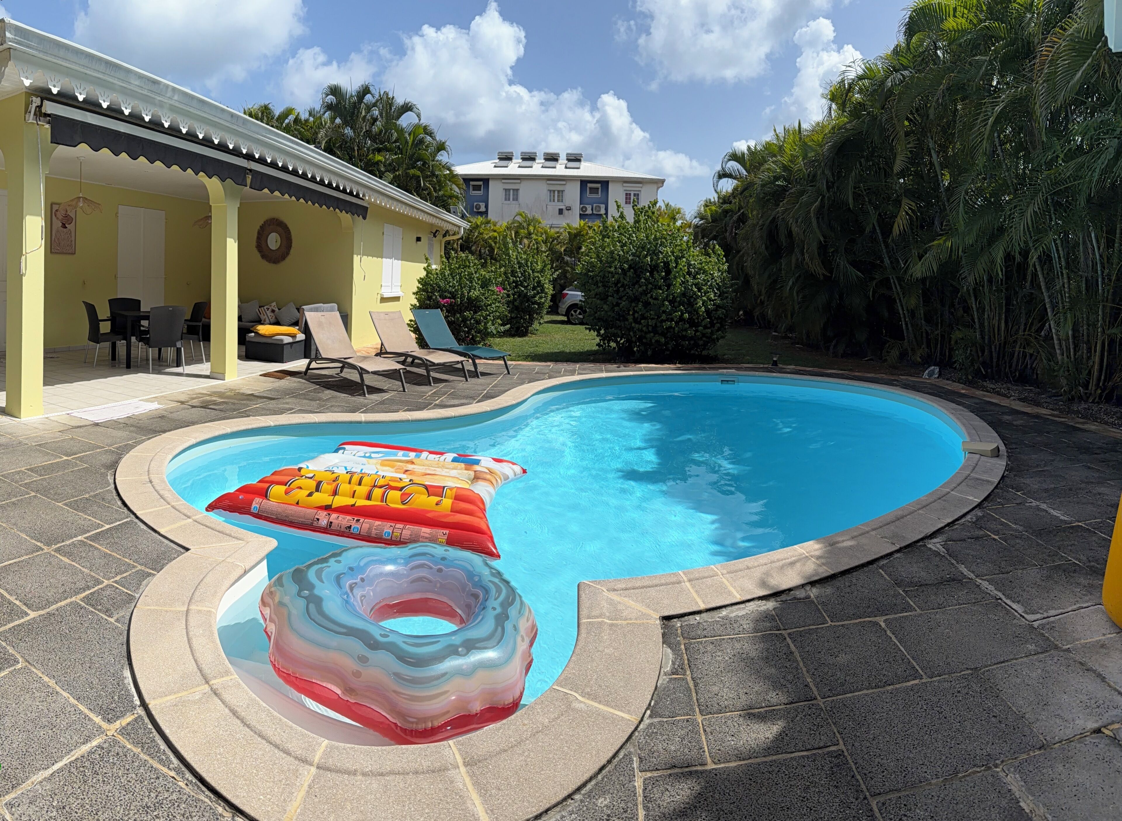 Une piscine avec deux bouées gonflables, un patio couvert avec des chaises, et un bâtiment en arrière-plan.