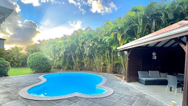 Ein Pool mit ungewöhnlicher Form, umgeben von Steinplatten, einer überdachten Außenlounge und üppigem Grün.