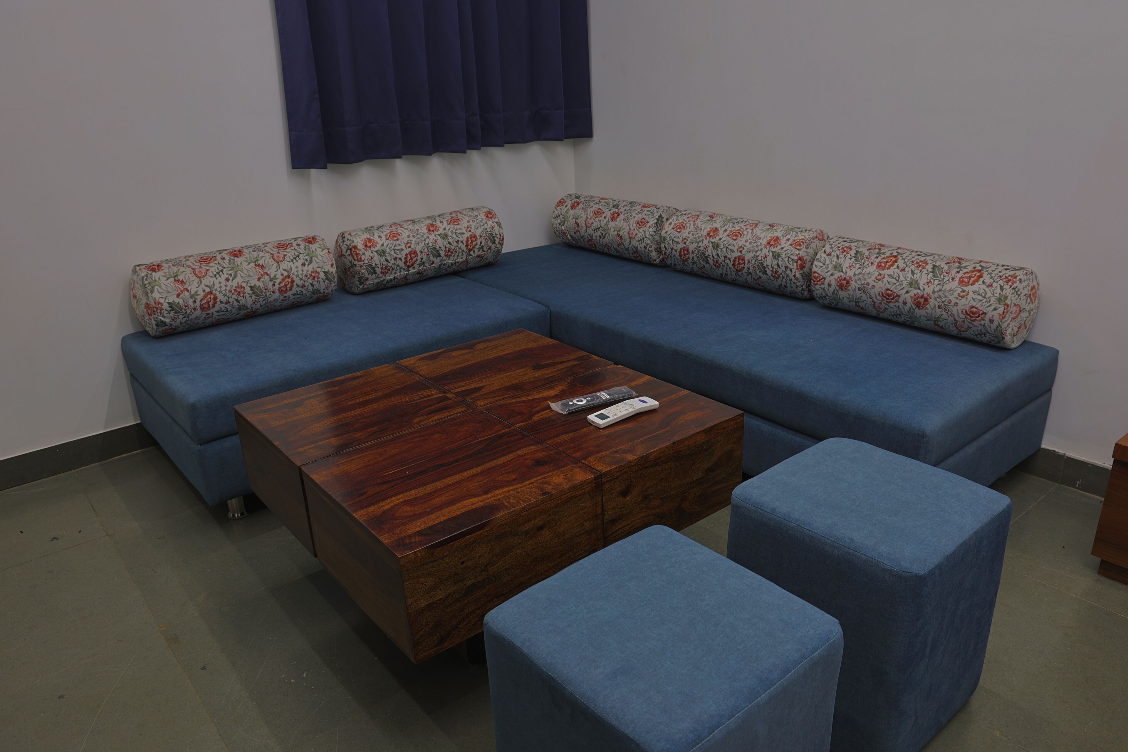 A blue sectional sofa with floral cushions, a wooden coffee table, and matching ottomans in a room with a plain white wall and dark curtains.