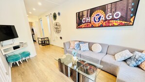 En moderne stue med sofa, sk footstols, et sofabord i glas og et fjernsyn. Et stort skilt med 'CHICAGO' pryder væggen.