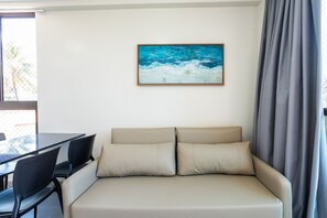Ein Zimmer mit einem beigen Sofa, zwei schwarzen Stühlen, einem Tisch und einem Wandbild von einem blauen Meer.