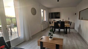 Quarto com mesa de jantar, cadeiras, televisão presa na parede, planta em vaso e vista através de uma porta de vidro.