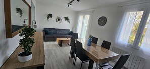 Un salon moderne comprenant un canapé, une table à manger, des chaises, une table basse, des plantes en pot, un miroir, des plantes fixées au mur, une horloge et de grandes fenêtres.