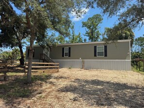 A single-story building with a covered porch, a fenced yard, and trees surrounding the property. - Happy Shores A Quaint 2-bedroom mobile home in Steinhatchee (Steinhatchee)