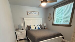 A bedroom with a bed, a nightstand, a ceiling fan, a window with blinds, and a welcome sign. - New 1-bedroom house in Garfield Bay perfect for a relaxing getaway (Sagle)
