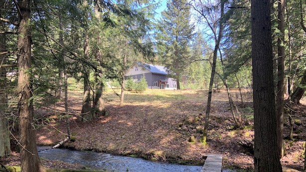 A forested area with a small stream, a wooden bridge, and a house in the background. - New 1-bedroom house in Garfield Bay perfect for a relaxing getaway (Sagle)