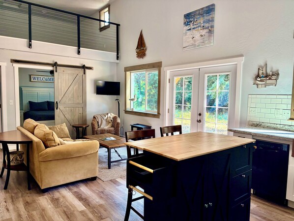 A modern kitchen with a central island, a dining area with a sofa and chairs, a TV mounted on the wall, and a view of the outdoors through large windows.