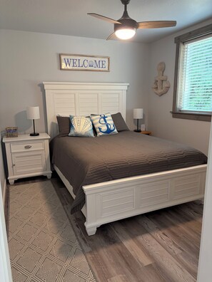 A bedroom with a bed, nightstands, a ceiling fan, a welcome sign, and a window with blinds. - New 1-bedroom house in Garfield Bay perfect for a relaxing getaway (Sagle)
