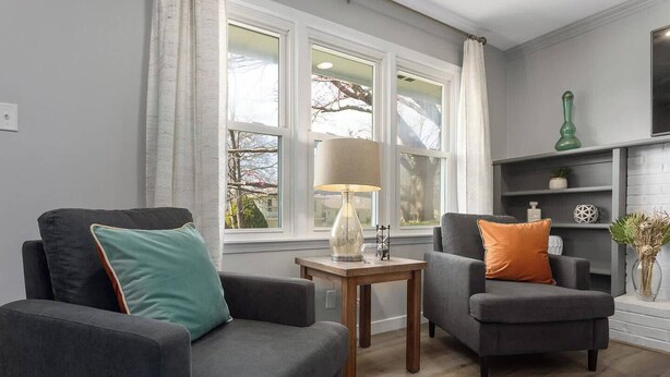 Ett vardagsrum med två fåtöljer, ett litet bord med en lampa och en öppen spis med dekorativa föremål.