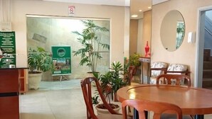 En hotellobby med en receptionsdisk, trästolar, ett runt bord, krukväxter och en stor spegel.
