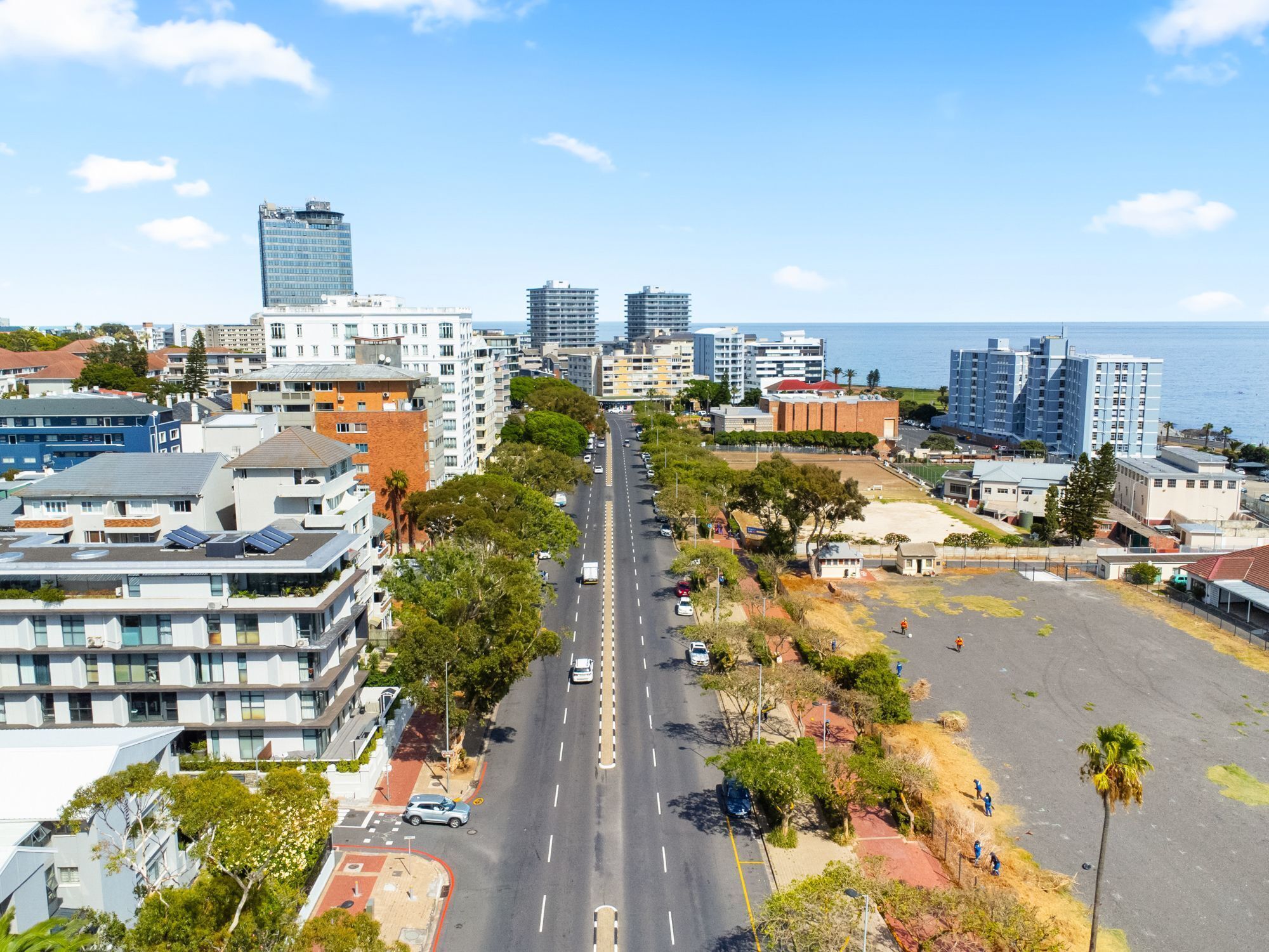 A cityscape with modern buildings, a wide road, and a clear view of the sea.