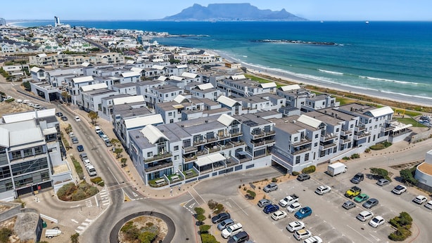 An aerial view of a coastal residential area with modern apartment buildings, a road, and a beach with a mountain in the background. - Eden On The Bay 231 - Two Bedroom Apartment, Sleeps 4 (Cape Town)