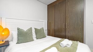 A neatly made bed with white linens and green pillows, a bedside lamp, and a wooden wardrobe. - Eden On The Bay 231 - Two Bedroom Apartment, Sleeps 4 (Cape Town)