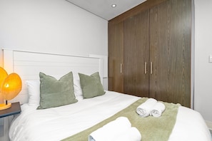 A neatly made bed with white linens and green pillows, a bedside lamp, and a wooden wardrobe. - Eden On The Bay 231 - Two Bedroom Apartment, Sleeps 4 (Cape Town)