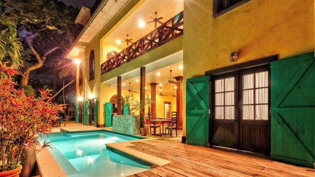 Area piscina con pavimento in legno, piscina e edificio con balcone e persiane verdi.