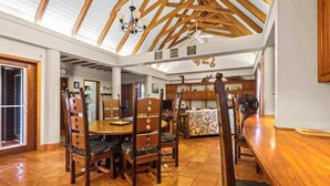 Uma sala de jantar espaçosa com teto alto e vigas de madeira, uma mesa central com cadeiras e vista para a área da cozinha.