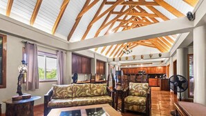Uma sala de estar ampla, com teto alto, vigas de madeira, um sofá, uma mesa de centro e uma cozinha ao fundo.