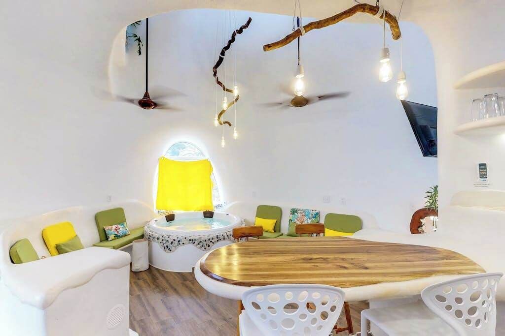 Un intérieur moderne avec une table ronde en bois, des canapés blancs ornés de coussins verts, une petite table ronde recouverte d’une nappe jaune et un ventilateur de plafond.