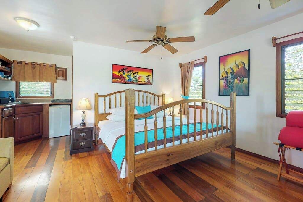 Una camera da letto con un letto di legno, una ventola a soffitto, un piccolo comodino, un tavolino di fianco al letto, una finestra con tende e un quadro appeso al muro.