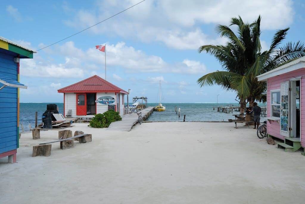 Ein Steg am Strand mit einem kleinen, roten Gebäude, auf dem 'Blue Wave Dive' steht, ein gelbes Boot liegt angedockt und es gibt einen pinkfarbenen Kiosk.