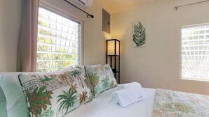 A hotel room with a bed, a window with blinds, a wall-mounted air conditioner, and a framed leaf artwork. - Colorful studio cabana with WiFi, partial AC, yard & picnic area - walk to town (Caye Caulker)