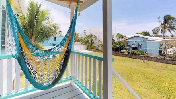 A hammock on a porch with a view of a residential area and a blue building. - Colorful studio cabana with WiFi, partial AC, yard & picnic area - walk to town (Caye Caulker)