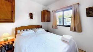 Un dormitorio con cama, mesita de noche con lámpara, ventana con cortinas y un armario de madera.