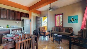 A dining area with wooden chairs and a table set for a meal, a living room with a sofa and armchairs, and a kitchen with cabinets and a refrigerator. - Inviting apartment w WiFi & partial AC - steps from the ocean, walk everywhere (San Pedro)