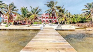 A wooden dock leading to a building with a red facade and white balconies, surrounded by palm trees. - Breezy oceanfront, 2-story condo w shared pool, beach access, WiFi & partial AC (San Pedro)