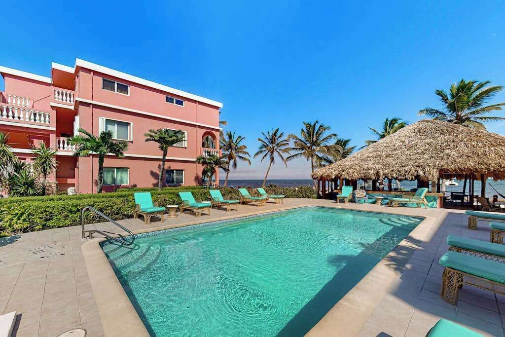 Un espace piscine avec des chaises longues, un parasol en chaume et un bâtiment doté de balcons.