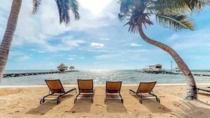 A beach with three sun loungers, a palm tree, and a thatched-roof structure in the distance. - Breezy oceanfront, 2-story condo w shared pool, beach access, WiFi & partial AC (San Pedro)