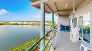A balcony with a hanging chair, a view of a calm body of water, and a distant bridge. - Seaside Waterfront Condo w Ocean View, Pool, Central AC & WiFi - Dogs OK (San Pedro)