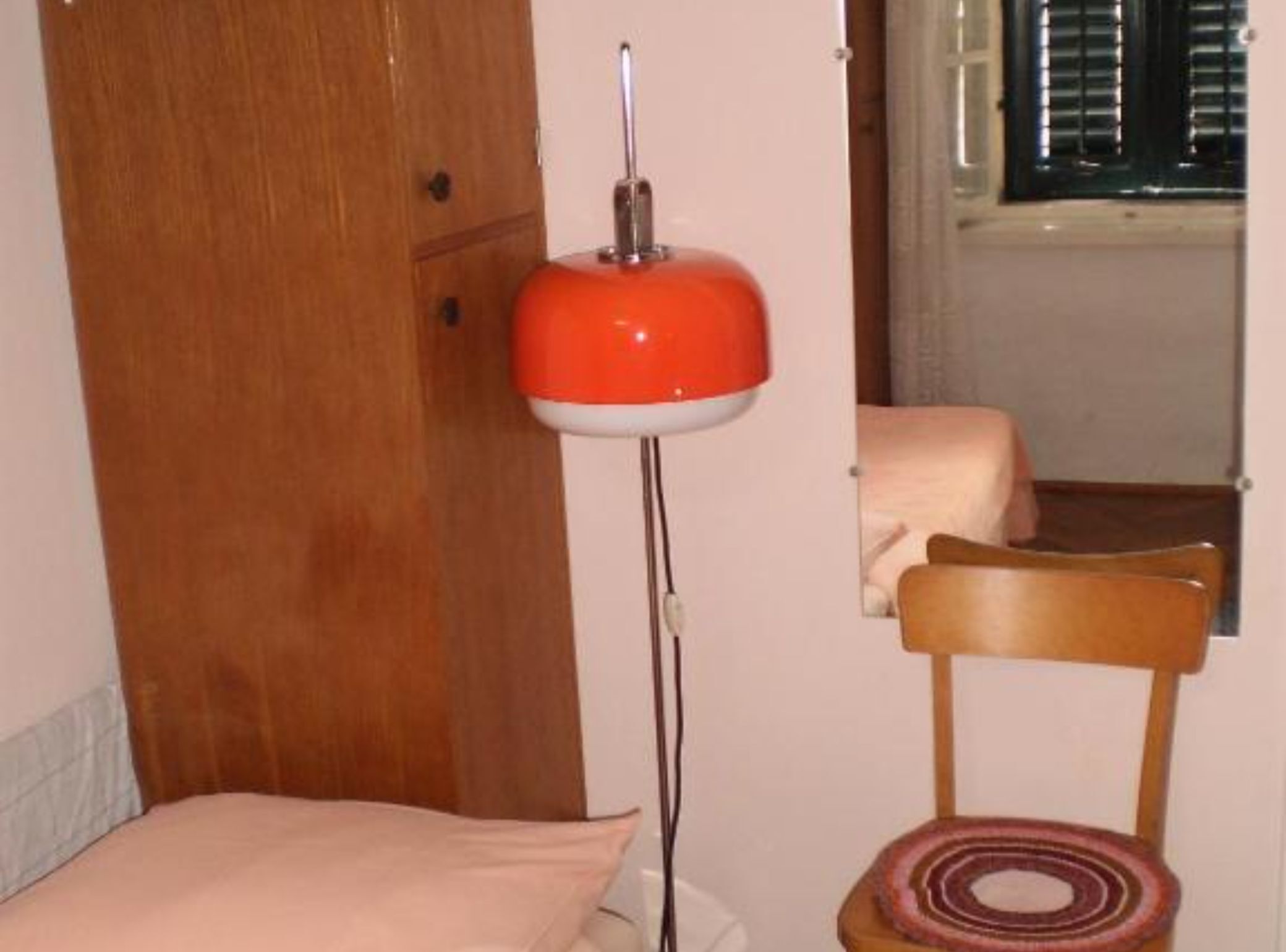 A room with a wooden door, a red floor lamp, a wooden chair, and a bed with a white pillow.