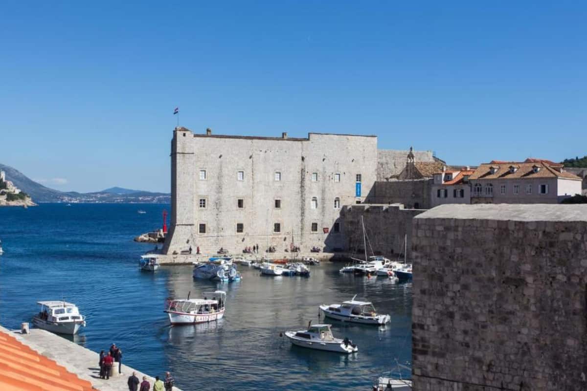 Un bâtiment historique en pierre, surmonté d’un drapeau, un port avec des bateaux et un ciel bleu azur.