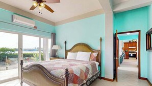 Ein Schlafzimmer mit Bett, Deckenventilator, Klimaanlage, Blick durch eine Glastür und einer sichtbaren Küche durch eine offene Tür.