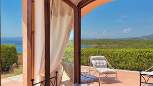 An open patio with a table and chairs, a view of the sea, and a lush green landscape. - Il Delfino - Seaside Bliss (Golfo Aranci)