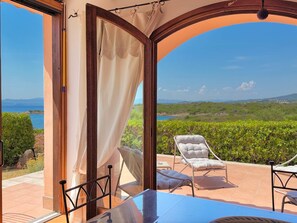An open patio with a table and chairs, a view of the sea, and a lush green landscape. - Il Delfino - Seaside Bliss (Golfo Aranci)