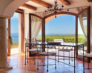 A room with a table and chairs, large windows with curtains, and a view of the sea and hills. - Il Delfino - Seaside Bliss (Golfo Aranci)