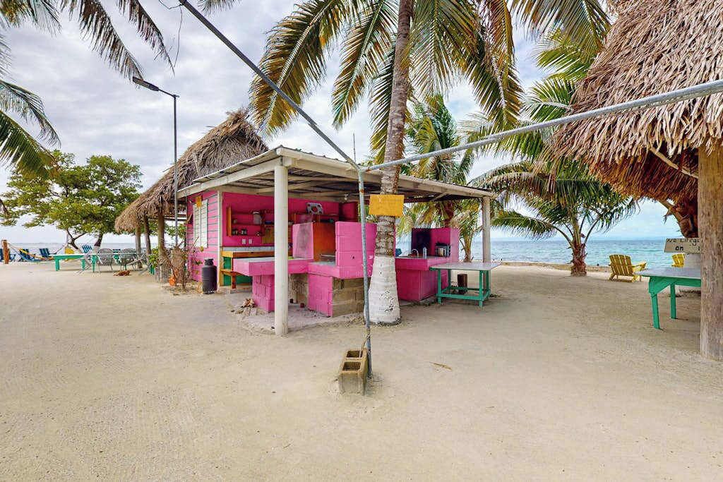 Un petit chalet au bord de la plage, avec un toit de chaume, une table et des chaises, entouré de palmiers et bordé par une plage de sable.
