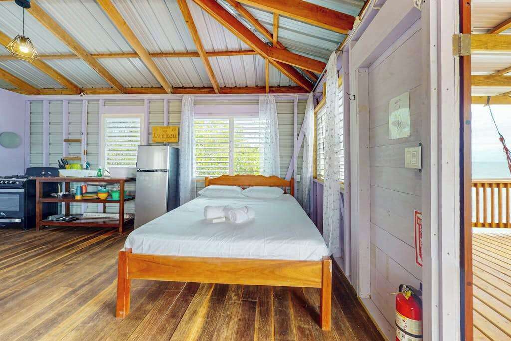 Una habitación con una cama de madera, un escritorio con un ordenador, un refrigerador y una ventana con persianas.