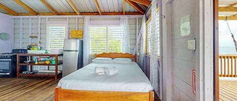 Una habitación con una cama de madera, un escritorio con un ordenador, un refrigerador y una ventana con persianas.