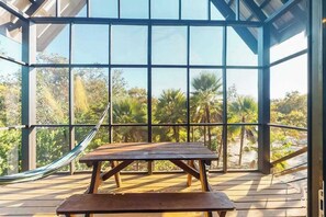 Een houten terras met een hangmat en picknicktafel, grote ramen en uitzicht op palmbomen en groen.