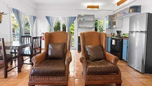 Una habitación con dos sillones de mimbre, una mesa de comedor, una zona de cocina con refrigerador y un ventilador de techo.