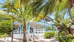 Un edificio de dos pisos con fachada blanca y detalles azules, rodeado de palmeras y una terraza de madera.