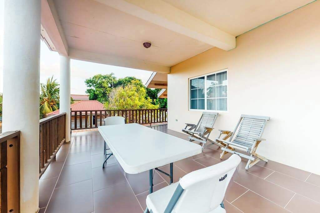 A covered patio area with a table and two lounge chairs, a window, and a view of greenery.