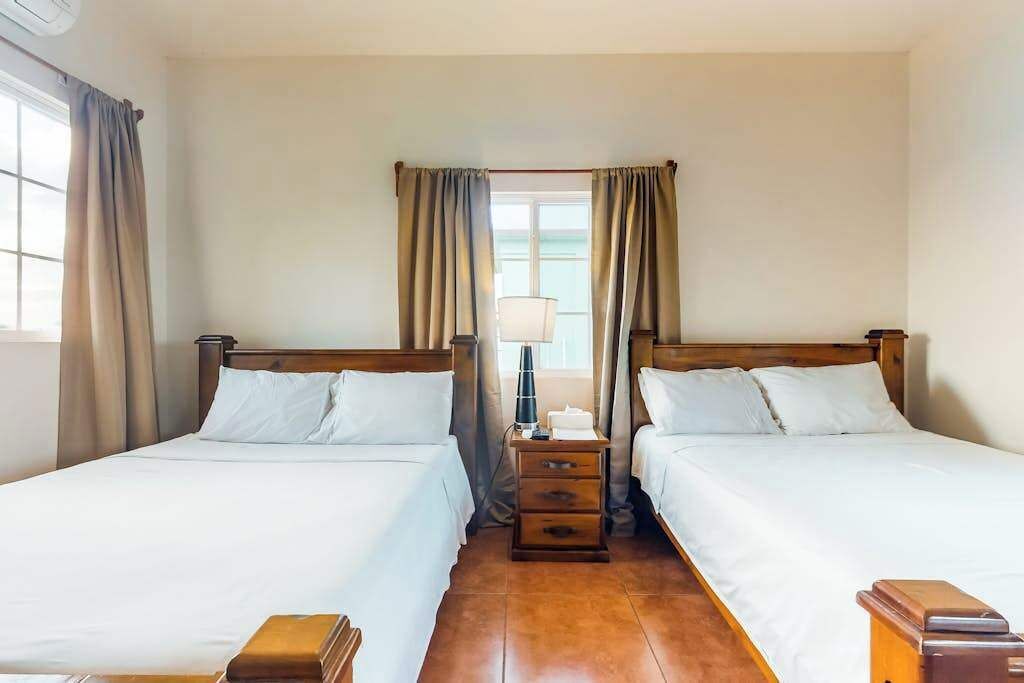 A hotel room with two beds, a nightstand, a lamp, and a window with curtains.