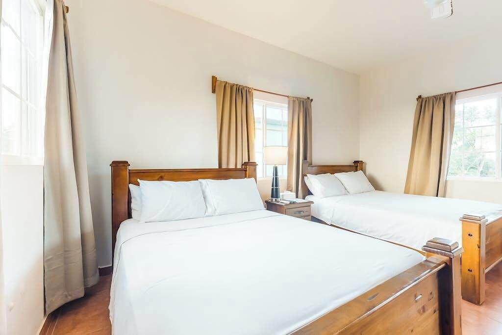 Habitación de hotel con dos camas, cabeceros de madera, ropa de cama blanca y cortinas.