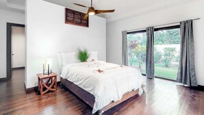 Ein Schlafzimmer mit einem Bett, einem Deckenventilator, einem Nachttisch, einem Fenster mit Vorhängen und Blick ins Grüne.