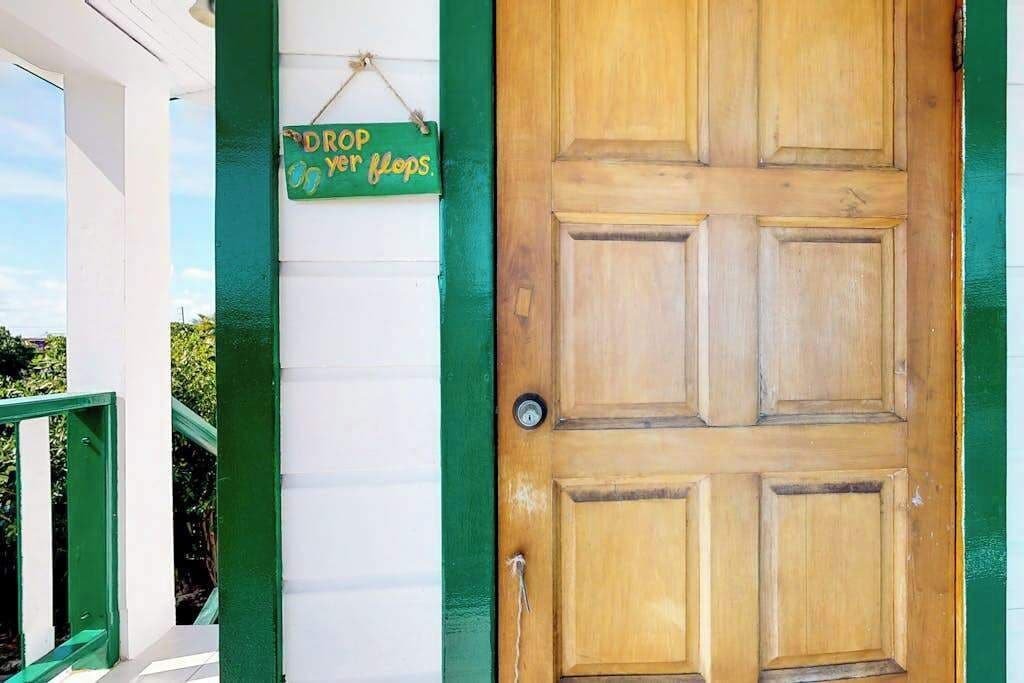 Une porte en bois avec un cadre vert, une enseigne suspendue sur laquelle on peut lire « DÉPOSEZ vos tongs », et un mur blanc avec une bordure verte.