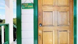 Une porte en bois avec un cadre vert, une enseigne suspendue sur laquelle on peut lire « DÉPOSEZ vos tongs », et un mur blanc avec une bordure verte.